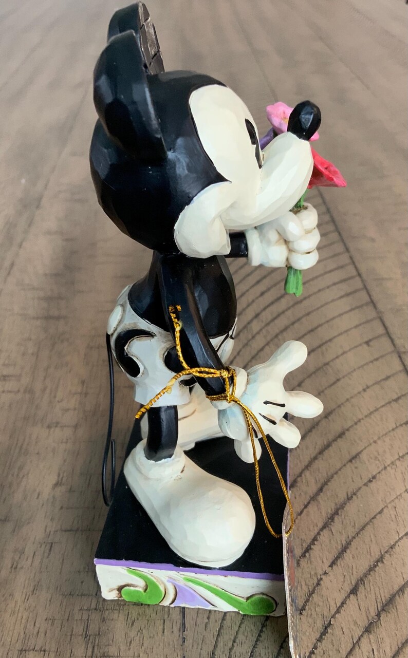 Op de afbeelding: Een Mickey Mouse-figuur met een uitstraling van gesneden hout, geschilderd in zwart, wit en cr&egrave;me. Het personage houdt een klein boeket van roze en rode bloemen vast. Een dunne gouden metalen draad is om de arm en taille gewikkeld.