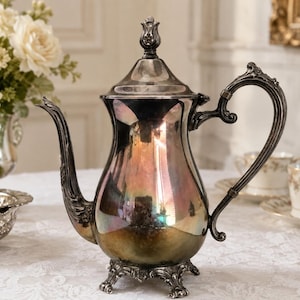 May include: A silver-plated teapot featuring a curved spout, an ornate handle, and a lid with a decorative finial. The body of the teapot has a polished, reflective surface with visible iridescent tarnish and stands on four detailed, scrolled feet.