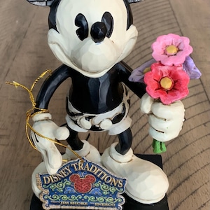 Op de afbeelding: Een Mickey Mouse-figuur met een zwart-wit lichaam en grote oren met een zwart-grijs ruitpatroon. Het personage houdt een boeket roze en paarse bloemen vast. Een Disney Traditions Jim Shore Enesco-label is aan de figuur bevestigd.
