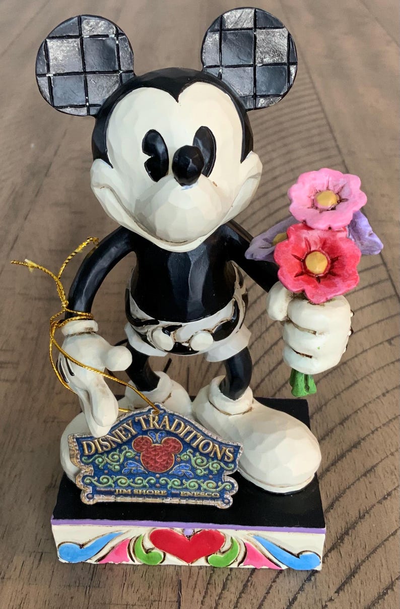 Op de afbeelding: Een Mickey Mouse-figuur met een zwart-wit lichaam en grote oren met een zwart-grijs ruitpatroon. Het personage houdt een boeket roze en paarse bloemen vast. Een Disney Traditions Jim Shore Enesco-label is aan de figuur bevestigd.