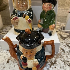 May include: A ceramic teapot and two figurines. The teapot is brown with a black lid and a face design. The figurines depict people in period clothing, one holding a basket and the other seated. The items are on a white surface.