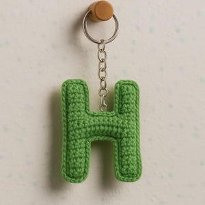 Peut inclure: Un porte-clés en forme de lettre "H" crochetée verte. La lettre est attachée à une chaîne et un porte-clés argentés, et est suspendue à une cheville en bois. Le porte-clés est fait d'un fil vert texturé.