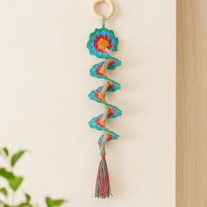 May include: A handmade crochet spiral decoration in a vibrant array of colours. The spiral design features blue, green, orange, and red, with a grey central section. It is suspended from a wooden ring and has a tassel. The spiral is approximately 50 cm long.