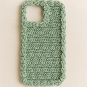 Può includere: Una custodia per telefono all'uncinetto verde salvia. La custodia presenta un motivo a punto strutturato e un bordo decorativo di punti a nocciolina. L'apertura per la fotocamera è visibile nella parte superiore. La custodia è progettata per proteggere un telefono.