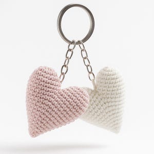 May include: A keychain featuring two crocheted hearts, one in pale pink and the other in white, attached to a silver chain and ring. The hearts are made of textured yarn, set against a white backdrop.