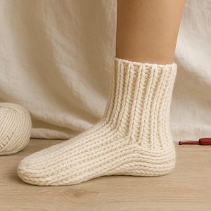 Puede incluir: Calcetín de punto grueso color crema. El calcetín es acanalado y parece hecho a mano. Se ve una madeja de lana en el fondo. El calcetín está en un pie, lo que sugiere un diseño cálido y acogedor.