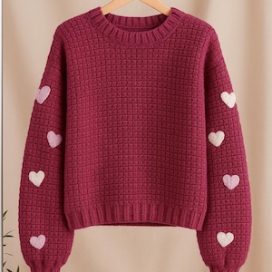 May include: A burgundy crew neck jumper with a textured knit pattern. The sleeves are decorated with white and light pink heart appliqués. The jumper is hanging on a wooden hanger.