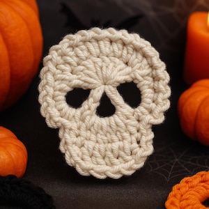 May include: A cream-coloured, crocheted skull-shaped coaster. The coaster has two eye sockets and a nose cavity. The skull is surrounded by pumpkins and a lit candle, suggesting a Halloween theme.