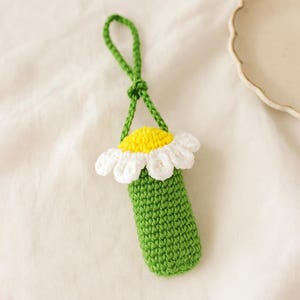 Pode incluir: Um porta-chaves de margarida de crochê feito à mão. O porta-chaves apresenta um corpo cilíndrico verde, uma flor de margarida branca e amarela e uma alça verde para pendurar. A margarida tem pétalas brancas e um centro amarelo. O porta-chaves é feito de fio.