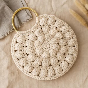 Puede incluir: Bolso de mano redondo de ganchillo color crema con asa dorada. El bolso presenta un patrón texturizado y en relieve de puntos de bobble. El asa tiene un diseño circular y rígido. El bolso está sobre un fondo neutro.