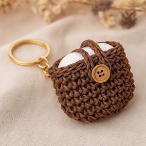 Può includere: Un portachiavi a forma di cestino all'uncinetto marrone con un anello dorato. Il cestino ha un manico e una chiusura con bottone in legno. Due oggetti bianchi sono visibili all'interno del cestino. Il portachiavi è su una superficie beige.