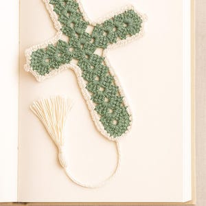 May include: A handmade crochet bookmark in the shape of a cross, featuring a green floral design with a cream-colored border. A cream-colored tassel hangs from the bottom. The bookmark rests on the open pages of a book.