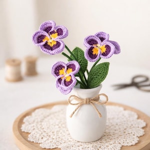 Op de afbeelding: Een kleine witte vaas bevat drie gehaakte viooltjes. De bloemen zijn paars met gele en donkerpaarse centra. Groene bladeren en stelen ondersteunen de bloemen. De vaas is vastgebonden met een touwstrik en staat op een houten dienblad met een kanten kleedje.