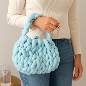 May include: A light blue, hand-braided handbag with a rounded shape and two braided handles. The bag is made of a soft, chunky material. The bag is held by a person wearing a white sweater and blue jeans.