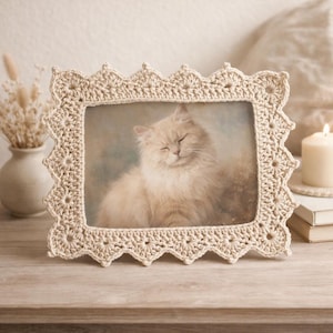 Puede incluir: Un marco de fotos beige tejido a ganchillo con un borde festoneado. El marco contiene una foto de un gato esponjoso de color crema con los ojos cerrados. El marco está sobre una superficie de madera, creando una estética acogedora y artesanal.