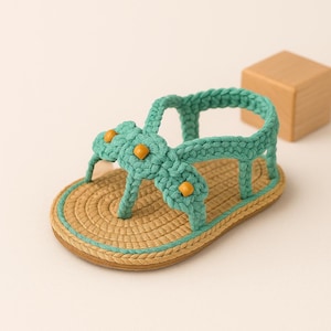 May include: A handmade crochet sandal in a light green hue, featuring a woven design with a tan sole and a light blue trim. The sandal has decorative yellow beads and a strap that goes around the ankle. The sandal is a small size.