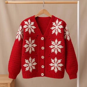 May include: A red, v-neck cardigan sweater with white snowflake designs. The sweater has a button-up front and is hanging on a wooden hanger. The snowflake patterns are on the front and sleeves.