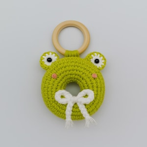 May include: A crocheted frog-shaped baby toy in lime green, with white and black eyes, pink cheeks, and a white bow. It features a wooden ring for grasping. A delightful and safe toy for infants, perfect for sensory play.