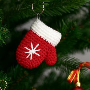 May include: A red and white crocheted Christmas mitten ornament with a white snowflake design. The mitten is hanging from a silver metal ring, and is displayed on a Christmas tree.