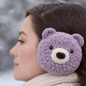May include: A close-up of a lavender-colored, crocheted bear ear warmer. The bear design features a white muzzle, a brown nose, and black eyes. The ear warmer is worn by a person with brown hair, and a cream-colored scarf.
