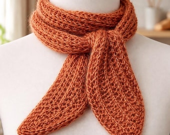 May include: An orange knitted scarf displayed on a mannequin. The scarf has a detailed open weave pattern. The scarf is tied in a knot, with the ends hanging down. The color is a warm, earthy orange. The scarf is made of a textured yarn.