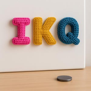 Może przedstawiać: Zbliżenie trzech szydełkowych liter, które tworzą napis "IKQ" w jasnych kolorach. Litera "I" jest w kolorze gorącego różu, "K" jest żółte, a "Q" jest turkusowe. Litery są przymocowane do białej powierzchni. Mały, okrągły, szary magnes leży na drewnianej powierzchni.