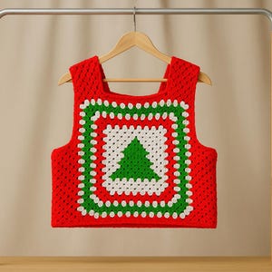 May include: A red, white, and green crochet vest with a Christmas tree design. The square-shaped vest is made of crocheted yarn and hangs on a wooden hanger. The Christmas tree is in the center of the vest.