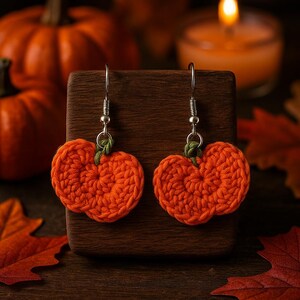 May include: A pair of orange crochet pumpkin earrings with silver hooks. The earrings are displayed on a wooden block, with pumpkins and autumn leaves in the background. A festive accessory for the autumn season.