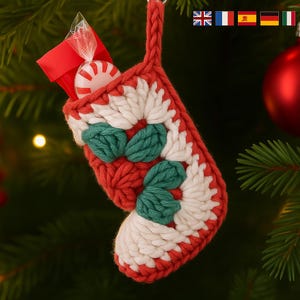 May include: A festive, handmade Christmas stocking in red, white, and green crochet, filled with a red gift bag and a peppermint candy. The stocking is decorated with white and green floral patterns and is hanging on a Christmas tree.