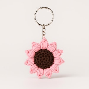 May include: A handmade keychain featuring a pink and brown sunflower design. The flower is made of yarn, with pink petals surrounding a brown center. The keychain has a silver metal ring and chain.