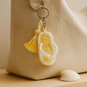 Può includere: Un portachiavi a forma di infradito giallo all'uncinetto con finiture e cinturini bianchi e un nappa gialla abbinata. Il portachiavi è attaccato a un anello color oro. Una conchiglia poggia su una superficie di legno.