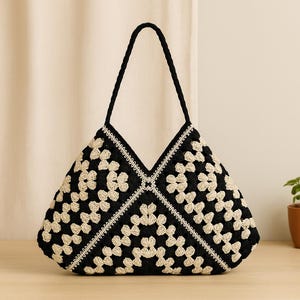 Op de afbeelding: Een zwarte en crèmekleurige gehaakte tote bag met een bloemenpatroon. De tas heeft een driehoekige vorm en een gevlochten zwart handvat. De randen zijn afgewerkt met witte stiksels. De tas staat op een licht houten oppervlak.