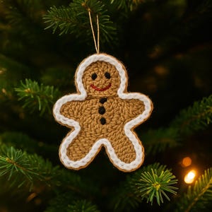 以下が含まれることがあります： 手作りのジンジャーブレッドマンのクリスマスオーナメント。茶色の糸でかぎ針編みされ、白で縁取られたオーナメントは、黒いボタンの目と赤い口を持つ笑顔の顔が特徴です。緑の松の針の背景に対して、金の紐から吊り下げられています。