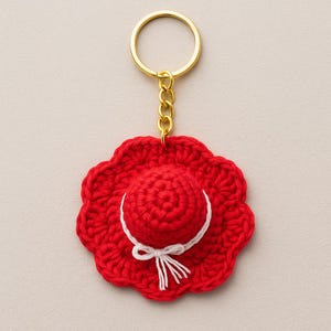Könnte beinhalten: Ein roter gehäkelter Hut-Schlüsselanhänger mit einem goldfarbenen Schlüsselring und einer Kette. Der Hut hat ein weißes Band und eine Schleife. Der Hut ist rund mit einem gewellten Rand.