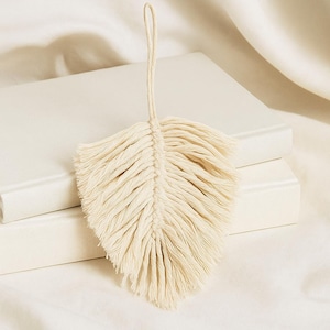 Può includere: Ornamento a forma di piuma in macramè color crema con un anello per appenderlo. Il design della piuma presenta uno stelo centrale con frange dettagliate, creando un'estetica strutturata e naturale. Ideale per l'arredamento bohémien.