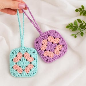 May include: Two handmade crochet ornaments. One is light blue with peach accents, the other is purple with peach accents. Each has a loop for hanging. The ornaments are square with a floral design in the center.