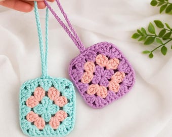 Puede incluir: Dos adornos hechos a mano con ganchillo. Uno es azul claro con detalles melocotón, el otro es morado con detalles melocotón. Cada uno tiene un lazo para colgar. Los adornos son cuadrados con un diseño floral en el centro.