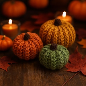 Pode incluir: Um conjunto de abóboras feitas à mão, tricotadas, em várias cores, incluindo laranja, amarelo e verde. Cada abóbora tem um caule castanho. As abóboras estão dispostas numa superfície de madeira com folhas de outono e velas acesas no fundo, criando uma cena acolhedora de outono.