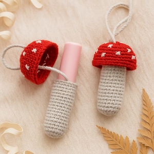 Puede incluir: Adornos de setas de ganchillo. Una seta tiene un gorro rojo con detalles de estrellas blancas y un tallo gris. Otra seta tiene un gorro rojo con detalles de estrellas blancas, un tallo gris y un tubo de pintalabios rosa. Las setas están hechas a mano y tienen una cuerda para colgar.