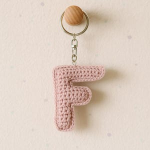 Può includere: Un portachiavi fatto a mano con la lettera "F" all'uncinetto rosa chiaro. La lettera è attaccata a un portachiavi argentato con un accento di perla di legno. Il portachiavi è appeso su uno sfondo rosa chiaro.