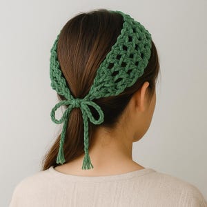May include: A green crochet headband with a bow at the back. The headband is made of thick yarn and has an open weave design. The bow has long, dangling ties with tassels at the ends. The headband is worn on a person's head.