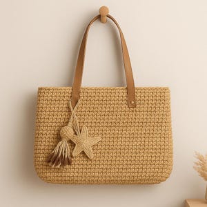 Könnte beinhalten: Eine gewebte beigefarbene Tragetasche mit braunen Ledergriffen hängt an einem Holzpflock. Ein dekorativer Quasten- und Seestern-Anhänger hängt an der Tasche. Die Tasche ist rechteckig und scheint handgefertigt zu sein.
