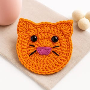 Op de afbeelding: Een handgemaakte oranje gehaakte kattenonderzetter met zwarte ogen, snorharen en een roze mond. De onderzetter is rond en heeft een kattengezichtontwerp. De onderzetter ligt op een beige servet.