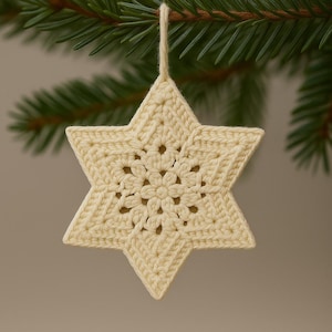 May include: A cream-colored, crocheted star ornament with a six-pointed design. The star is suspended by a matching string and hangs against a backdrop of green pine needles. The star's center features a floral pattern.