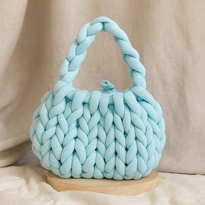 May include: A light blue, hand-knitted handbag with a braided handle. The bag is made of thick, soft yarn and sits on a small, round wooden base. The knitted texture is visible throughout the bag's body and handle.