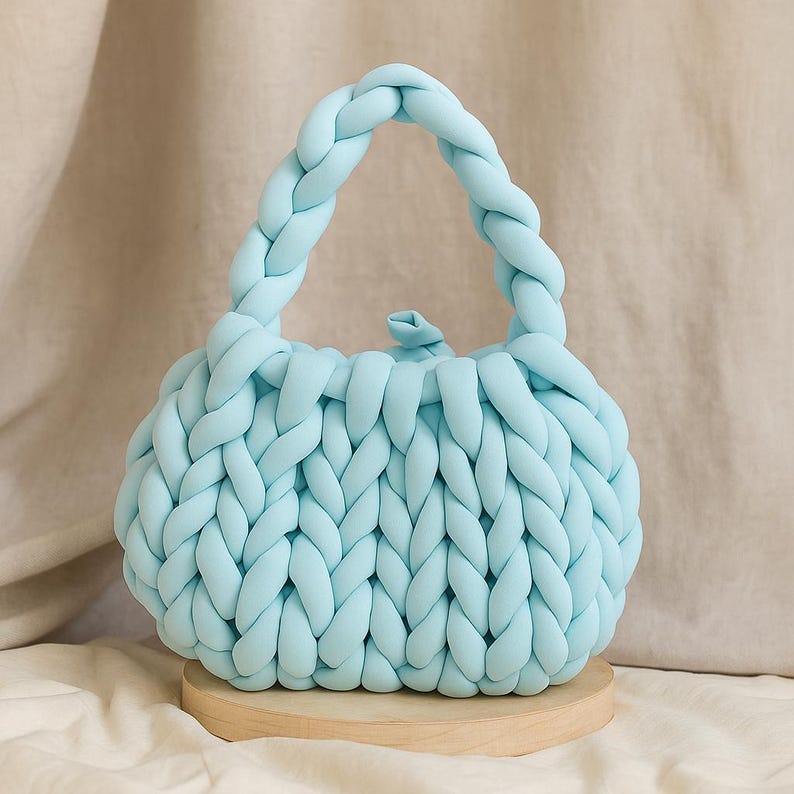 May include: A light blue, hand-knitted handbag with a braided handle. The bag is made of thick, soft yarn and sits on a small, round wooden base. The knitted texture is visible throughout the bag's body and handle.