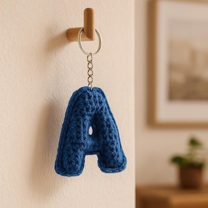 Puede incluir: Un llavero con la letra "A" azul tejido a ganchillo cuelga de un gancho de madera. El llavero está hecho de un hilo grueso y texturizado y está unido a un llavero y una cadena de metal. El fondo es una pared neutra con una imagen enmarcada.
