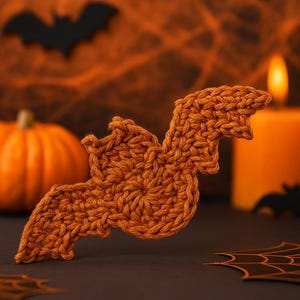 May include: A crocheted bat in shades of orange, with a textured surface. The bat is the focal point, set against a blurred background with a pumpkin, candle, and spiderweb, evoking a Halloween theme.