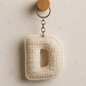 Puede incluir: Un llavero con la letra "D" de ganchillo, de color crema. La letra está unida a una cadena plateada y a una pieza circular de madera. El fondo es de color beige claro con lunares sutiles y coloridos.