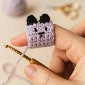 Op de afbeelding: Een handgemaakte haakring in de vorm van een berengezicht, met een lichtpaarse body en zwarte oren. De ring zit om een vinger, met een haaknaald en garen op de voorgrond. Een bol garen en een schaar staan op de achtergrond.